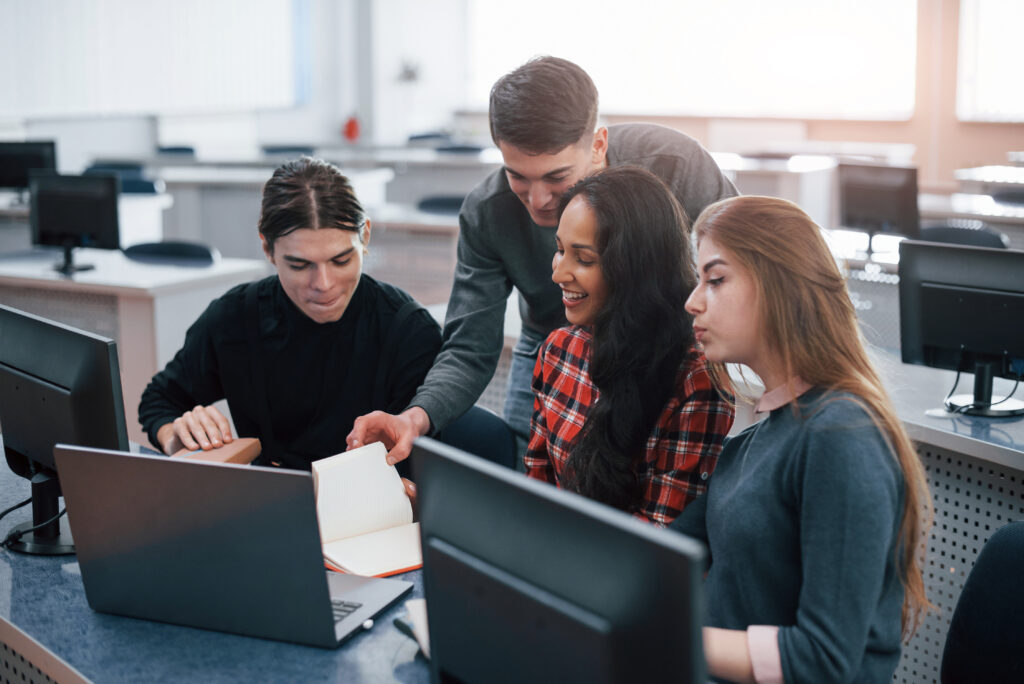 Bildungswesen haupt - Alt text: A group of young people wearing casual clothes are working together in a modern office. The group is standing around a large desk, looking at a computer screen and discussing their work. The office has large windows that let in natural light and there are plants and modern furniture in the background.