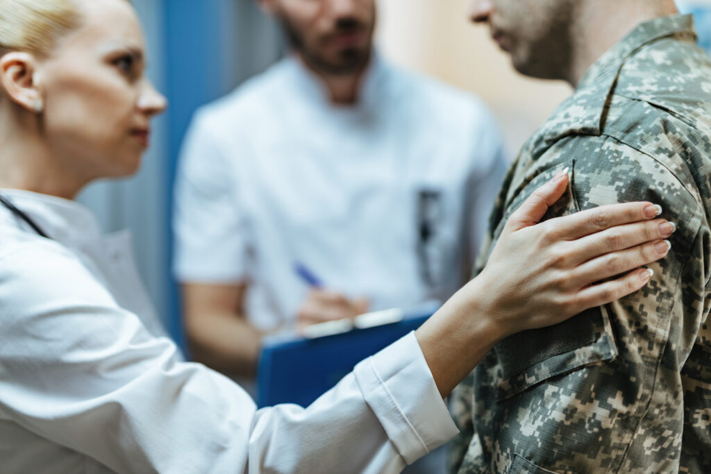 Militär - Alt text: A female doctor is shown in close-up, consoling a military man who is seated on a hospital bed. The man appears to be upset or in distress, and the doctor is placing a comforting hand on his shoulder while speaking with him. The hospital room is visible in the background, with medical equipment and a window with daylight coming in.